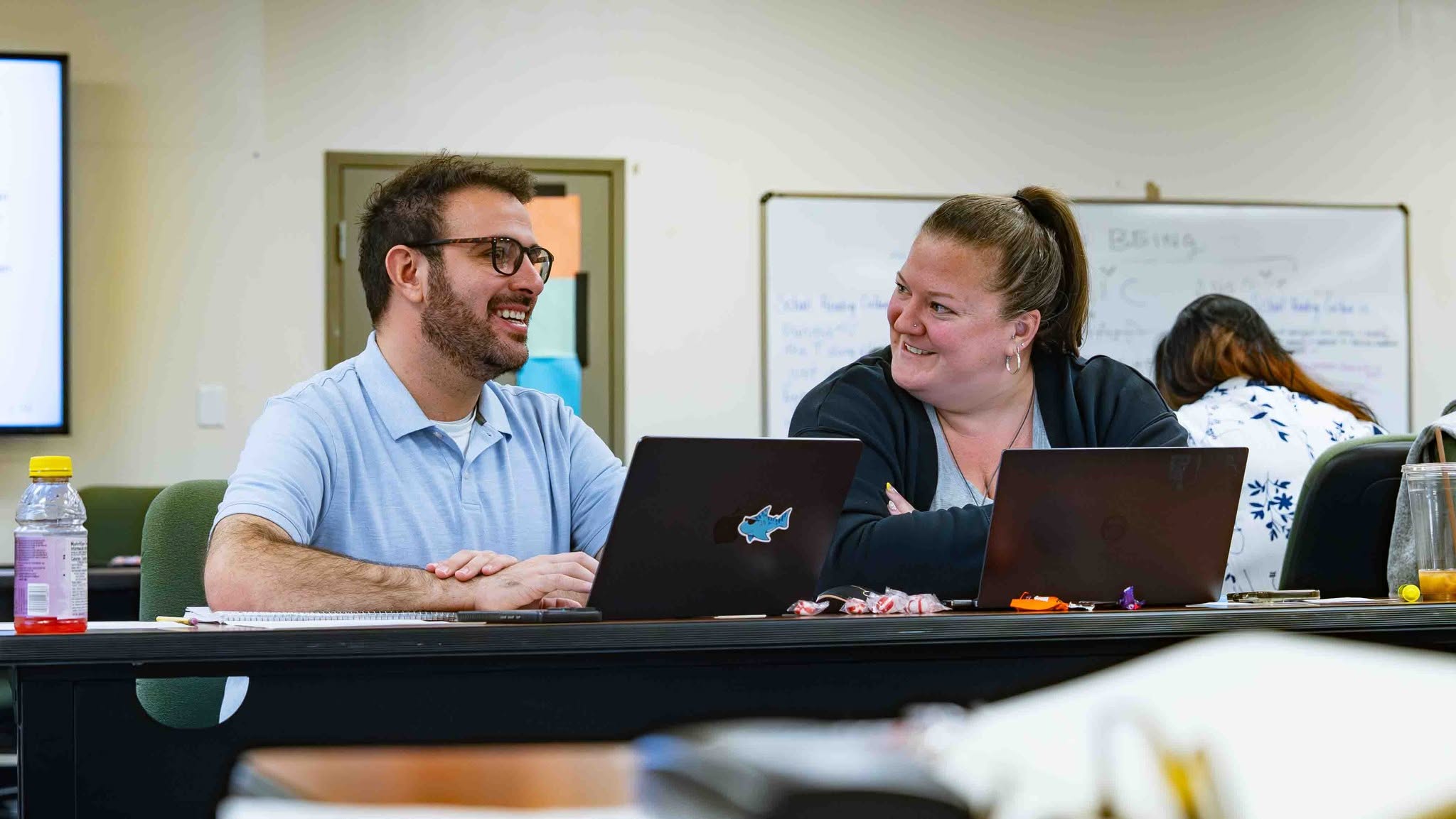 A male teacher and a female teacher are sitting at a table with their laptops open in front of them. The female teacher is turned and smiling at the male teacher while he smiles and looks at something off camera.