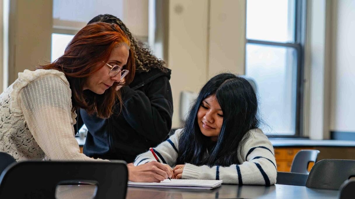 A female student is seated at a table with a notebook in front of her. A female educator is leaned over the notebook and writing on it.
